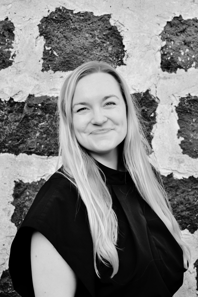 Black-and-white portrait of sara maaria toroi, who has long, light blonde hair and smiles softly at the camera. She wears a loose-fitting top that's pleated at the neckline and standing in front of a textured stone wall with large, irregular light and dark bricks.
