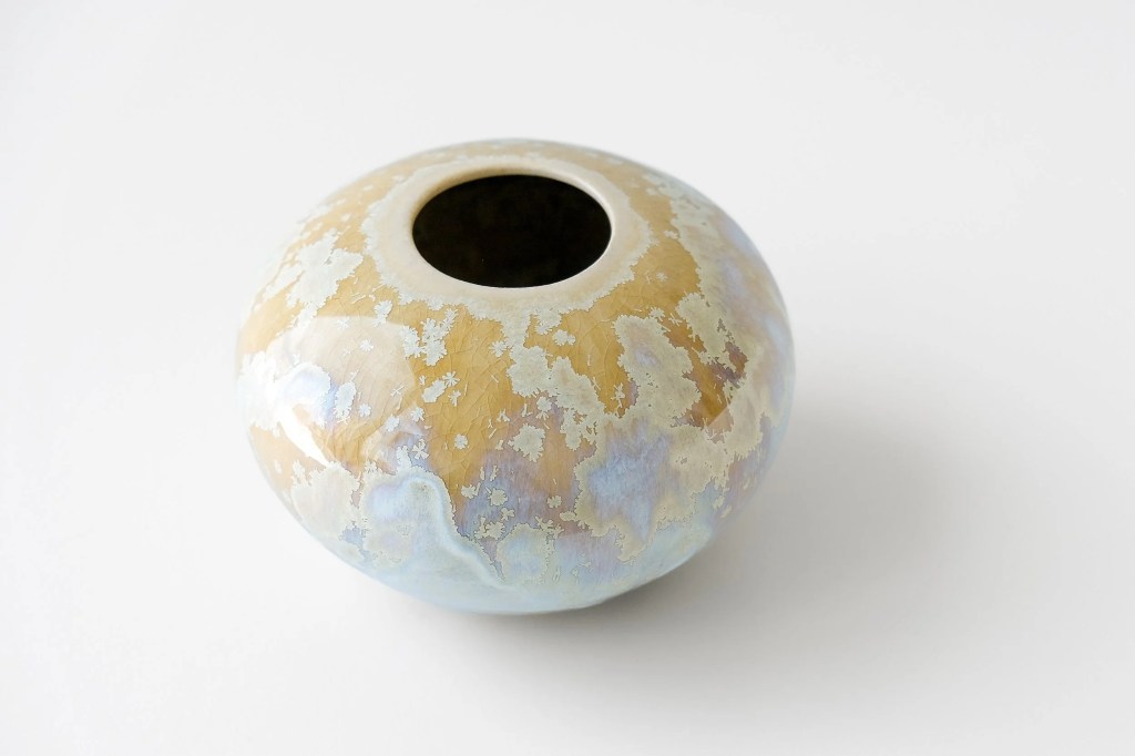 A rounded ceramic vessel with a circular opening at the top sits against a white background. The surface is a crackley crystalline glaze with light gold starbursts against a background of light ochre at the top and light blue and lavender hues at the base of the piece.